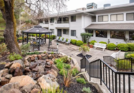 A white building with a patio and a black awning. The patio has a table and chairs, and a bench. There are potted plants and a tree in the area. AI generated content
